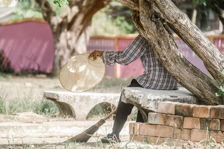 Old female cleaner sitting under the shadow of tree for rest and holding a sweep at the park.