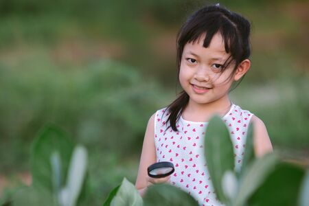Happy asian little child girl looking through a magnifying glass on green leaf tree in the parkの写真素材
