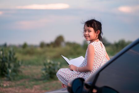 Little child girl  sitting and relaxing on a car reading a book in a green nature backgroundの写真素材