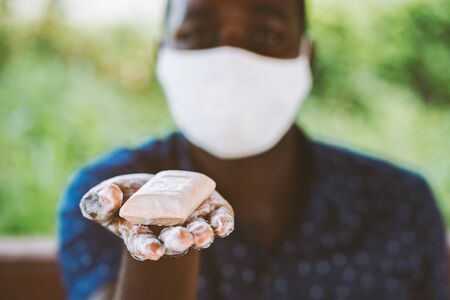 African man holds and show a piece of wet soap with wearing face maskの写真素材