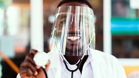 African doctor wear face shield and holding stethoscope with kind and smiling.16:9 styleの写真素材