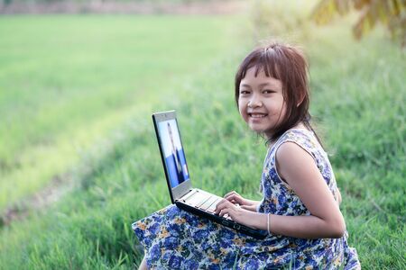 Happy little child girl learning outdoor by studying online and working on laptop in green fieldの写真素材