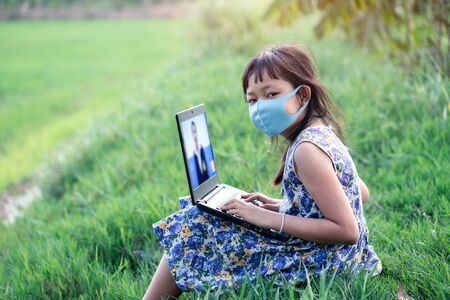 Happy little child girl learning outdoor by studying online and working on laptop in green field with wearing face maskの写真素材