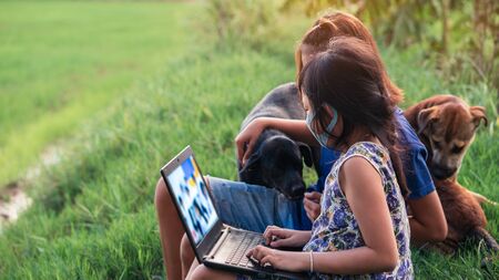 Happy two little child girl learning outdoor by studying online and working on laptop in green field with wearing face maskの写真素材
