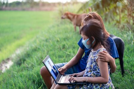Happy two little child girl learning outdoor by studying online and working on laptop in green field with wearing face maskの写真素材