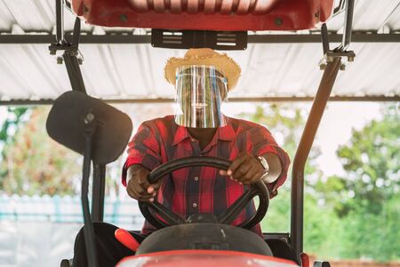 African farmer wear  face shield and driving tractor in farm during harvest in countryside. Agriculture or cultivation conceptの写真素材