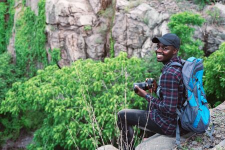 African man traveler sitting on the cliff and holding camera with backpackの写真素材