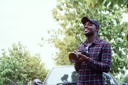 African man traveler is holding camera and standing near car freelancing on the roadの写真素材