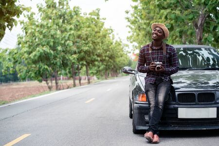 African man traveler is holding camera and standing near car freelancing on the roadの写真素材