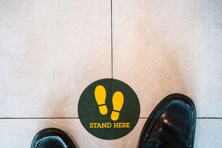 Black shoes standing on a green sign saying stand here to avoid spread of covid19の写真素材