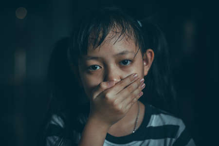 Sad little child girl covering her mouth with hands in dark roomの写真素材