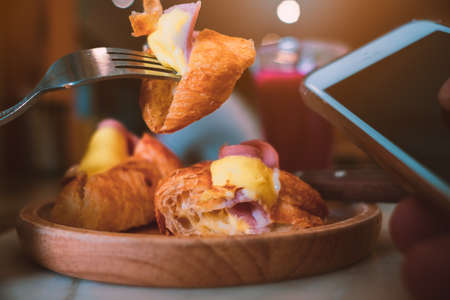 Person eating a piece of croissant by knife and fork in coffee shop with using smart phone