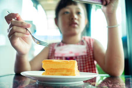 Asian little child girl eating chiffon cake with looking smart phoneの写真素材