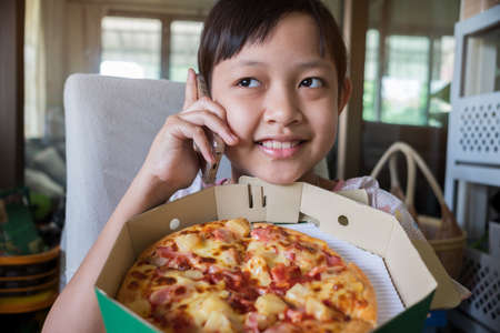 Asian little girl holding smartphone and ordering pizza to eat at homeの写真素材