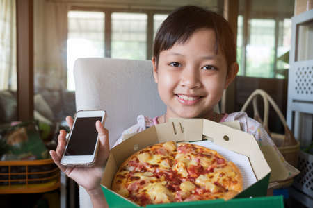 Asian little girl holding smartphone and ordering pizza to eat at homeの写真素材