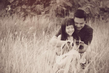 Kind asian father sitting on grass with little daughter sitting on him and reading a book.Vintage color styleの写真素材
