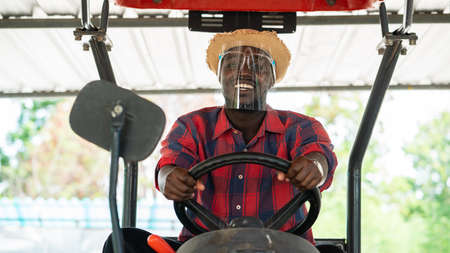 African farmer wear  face shield and driving tractor in farm during harvest in countryside. Agriculture or cultivation conceptの写真素材