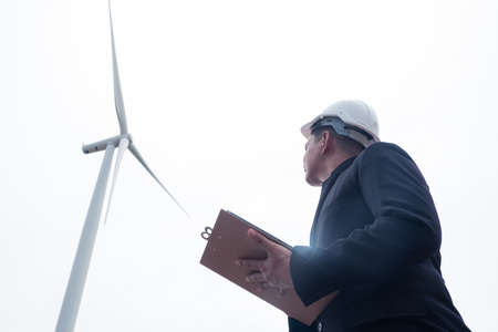 Male engineer looking and writing plan on clipboard against wind turbineの写真素材