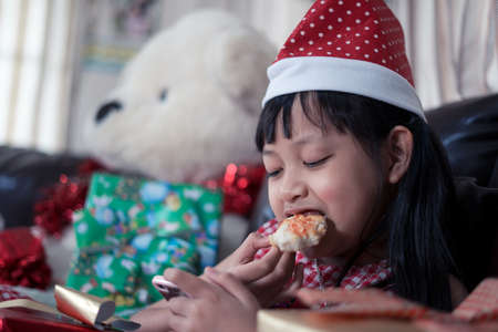 Happy asian child girl eating pizza and using smartphone in the room decorated for the new year. Christmas dinner fast foodの写真素材