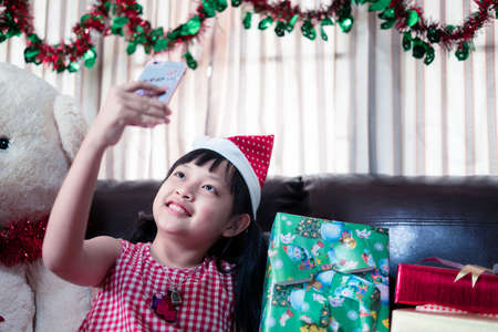 Asian little girl in a xmas Santa Claus hat looks at a smiart phone for video callの写真素材