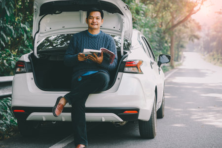 Asian traveler man is sitting and reading a book with open car trunkの写真素材