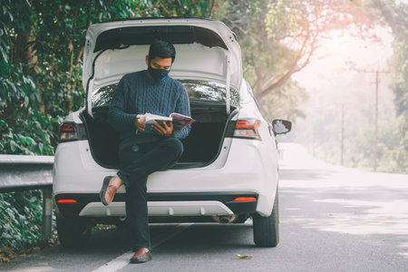 Asian traveler man wearing face mask and reading a book with open car trunkの写真素材