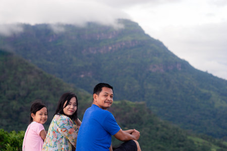 Happy asian family father mother and daughter sitting on top of beautiful mountain holding raised handsの写真素材
