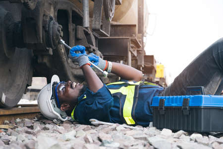 African machine engineer technician lying down and wearing a helmet, groves and safety vest is using...