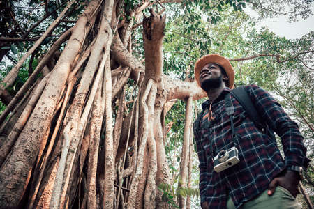 African man walking in the forest with a camera looking by side