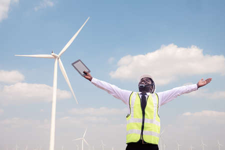 Successful african engineer windmills standing and hoding tablet with wind turbine in backgroundの写真素材