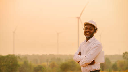 African engineer windmills wearing white hard hat and using tablet with   wind turbine on silhouette sunsetの写真素材