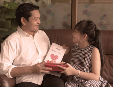 Asian little child daughters congratulating dad and giving him postcard and gift box with smiling and hugging.Concept of Happy father's dayの写真素材