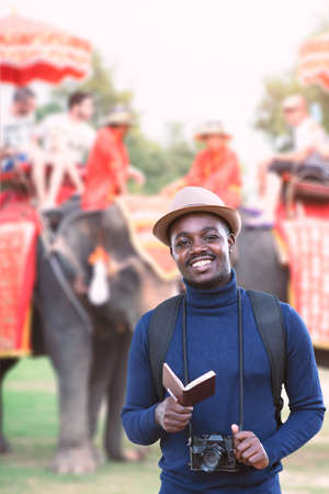 African traveler man travel in Thailand.He is holding passport and vintage camera with tourists on an ride elephantの写真素材