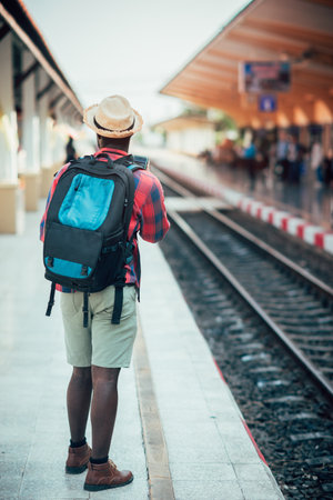 African male traveler with hat and backpack using smartphone and waiting for the train on railway station.Adventure travel conceptの写真素材