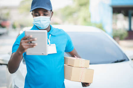 African delivery man wearing face mask and looking tablet with holding a box package .Concept of quarantine delivery serviceの写真素材