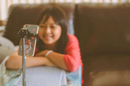 Asian little girl studying online at home, looking at phone screen with smile and happyの写真素材