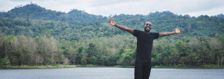 African man standing with arms outstretched in nature mountains and lake with smiles and happinessの写真素材