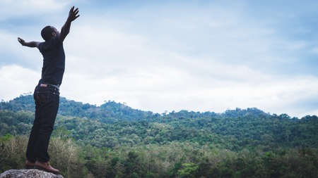 African man standing on the top of mountain with arms outstretched in nature and lake with smiles...