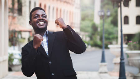 Excited african businessman keeping arms raised and expressing positivity while standing outdoors...
