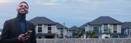 African smiling businessman stands in front of a housing estate while using a smartphone.Concept of Realty businessの写真素材