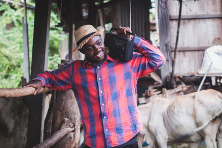 African farmer man with retro radio broadcast receiver on shoulder stands happy smiling outdoor on old cow stall backgroundの写真素材