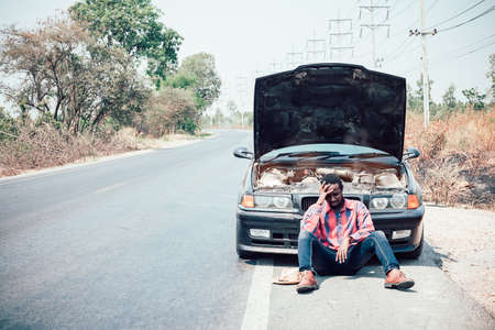 African man sits with his hand over his head as his car breaks down on the roadの写真素材