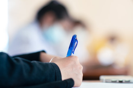 Student taking exam while wearing face mask due to coronavirus emergency. Young woman sitting in class with wearing surgical mask due to Covid-19 pandemicの写真素材