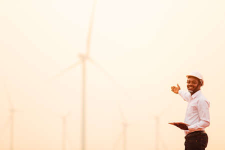 African engineer man stands holding tablet front the wind turbines generating electricity power station in sunset. 
Concept of sustainability development by alternative energyの写真素材