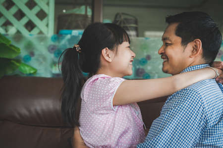 Father and daughter are hugging with love, looking at camera and smiling while sitting on sofa at home.Concept of Father's dayの写真素材