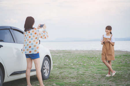 Happy mother and daughter taking photo by smartphone and having fun on a tropical beach and a car on the side. Family vacation holiday and travel conceptの写真素材