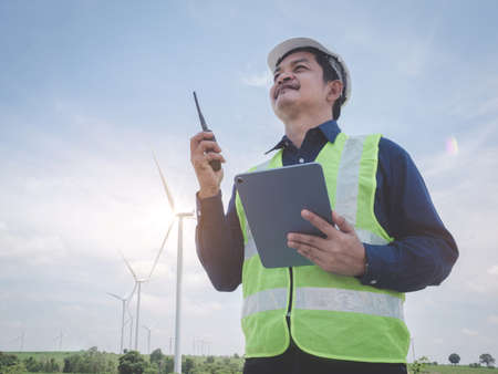 Asian engineer man stand holding tablet front the wind turbines generating electricity power station with talking by walkie -talkie . Concept of sustainability development by alternative energyの写真素材