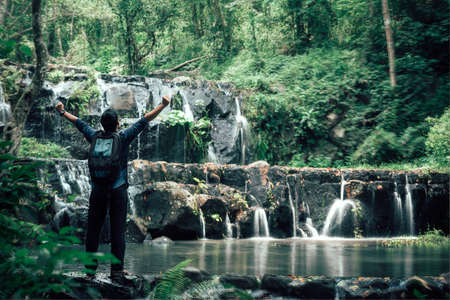 Freedom traveler man with backpack enjoying amazing tropical waterfall raising hands travel lifestyle and success vacation concept enter nature forest mountain backgroundの写真素材