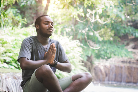 African man wearing a T-shirt sits in a waterfall to meditate and do yoga exercises