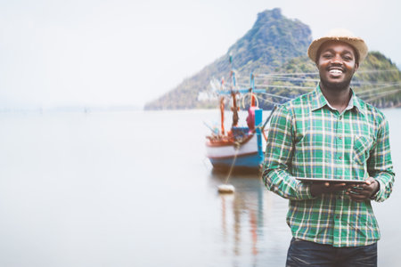 African trawler captain stands in front of the ship using a tablet to search for weather and maritime information.Marine business concepts and technologyの写真素材